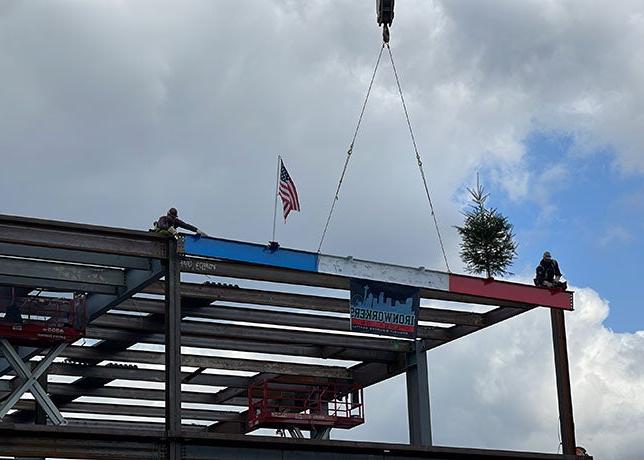 2 people on top of a steel structure on each side of a red, white and blue beam that has an american flag and an evergreen tree on it
